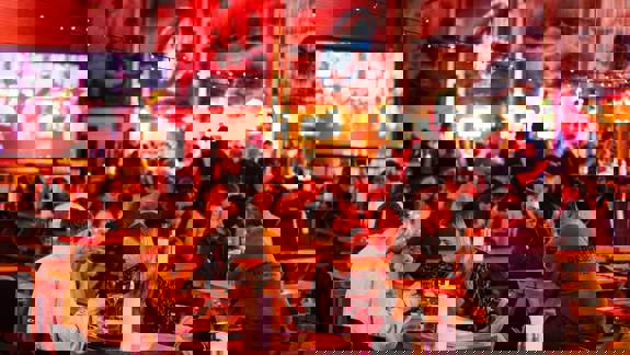 A wide shot of a bustling food market with long wooden tables and string lights hanging from the ceiling.