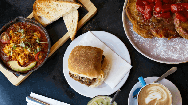 Brunch spread with shakshuka in skillet, avocado, toast, sandwich, pancakes with strawberries, cappuccino with latte art, iced matcha latte, and silverware on dark table.