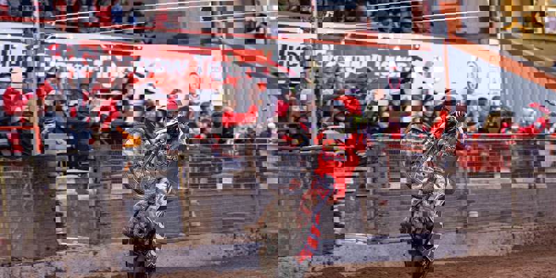 A dirt bike rider in red gear performs a wheelie on a dirt track, with cheering spectators in the background.