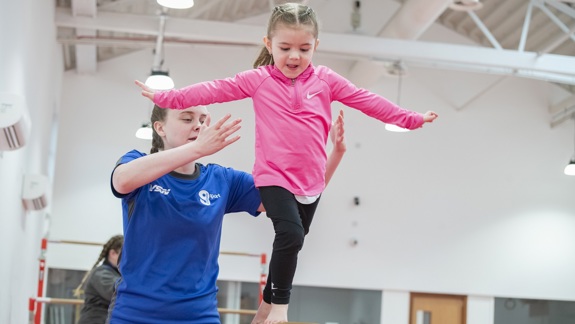 A young girl is balancing on a wooden balance beam with her arms out, with a female coach in a blue shirt standing beside her, ready to spot her.