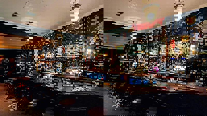  A warm, dimly lit traditional pub interior with a dark wood bar, bottles, glasses, and a Scottish flag display.