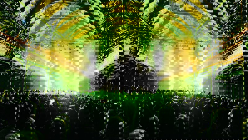 A crowded music venue with a band performing on a stage under bright spotlights, with the audience silhouetted in the foreground.