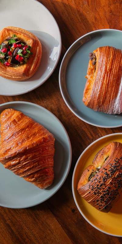 A wooden table holds four plates with pastries: a savoury danish with pomegranate, a chocolate pastry, a classic croissant, and a bun topped with seeds.