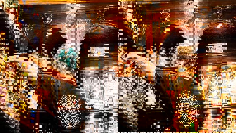 Bartender selects whisky bottles from a large, ornate shelf at The Pot Still, a traditional Glasgow whisky bar with a vast selection.