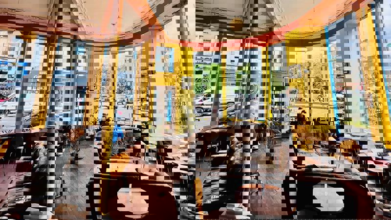 A cosy cafe interior with warm lighting, comfortable seating, and large windows looking out onto a street with cars and buildings.