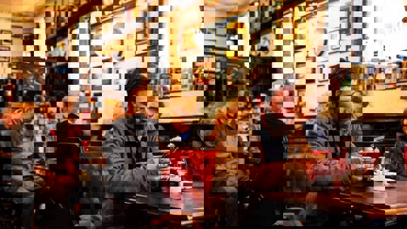 Customers relax inside The Pot Still, chatting or using phones in a warmly lit pub with red leather seating, framed decor, and whisky glasses on tables.