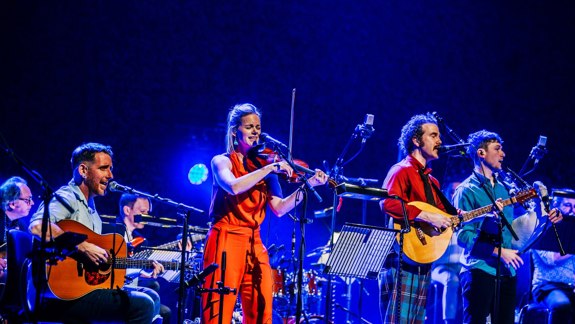 A music group performing on stage, featuring a woman playing a violin, a man playing a guitar, and another playing a bouzouki.