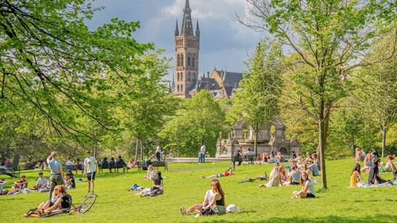 Kelvingrove Park: A park with people relaxing on the grass on a sunny day, with the University of Glasgow building visible in the background.