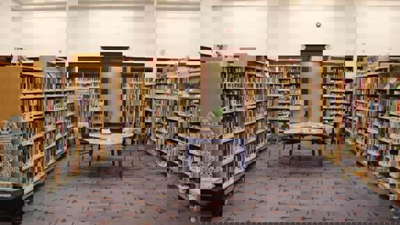 A wide shot of the Glasgow Women's Library reading room, featuring bookshelves filled with books, a round table with chairs, and patterned carpeting