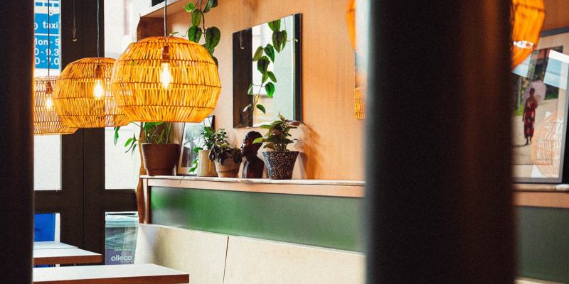 Wicker pendant lights hang above wooden tables in a cosy restaurant with plants on a shelf. A framed photo show a person walking.