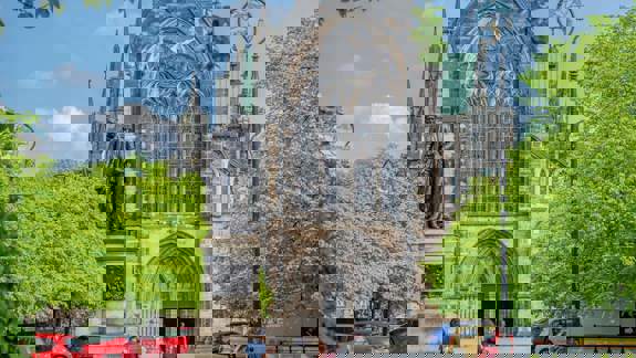 A full exterior shot of Glasgow Cathedral in Scotland. Lush green trees and parked cars surround the ancient stone building under a bright blue sky.