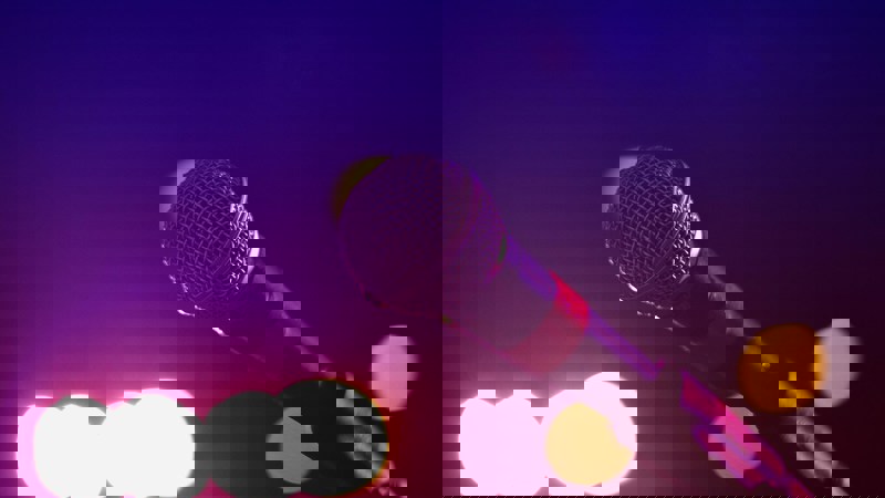 Close-up of a microphone on a stand under purple and pink stage lighting, with blurred circular lights in the background suggesting a live performance setting.