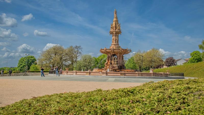 A ornate terracotta fountain, the Doulton Fountain, sits in a plaza with trees and people in the background under a blue sky.