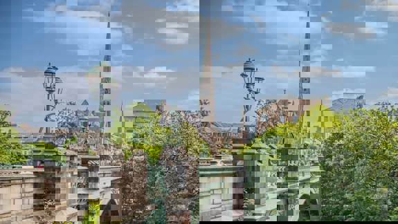 A bridge with decorative lampposts and a church spire in the background, surrounded by lush green trees.