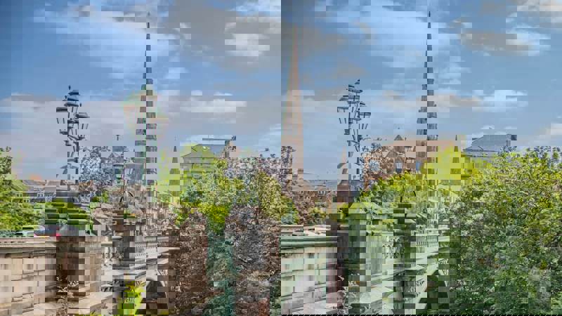 A bridge with decorative lampposts and a church spire in the background, surrounded by lush green trees.