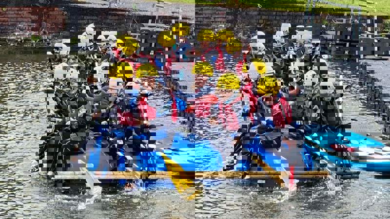 A group of children in yellow helmets and black wetsuits paddle a raft on the water.