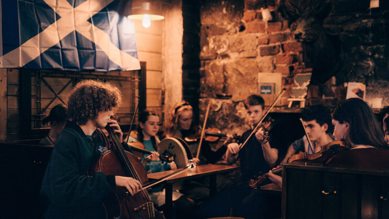 A group of young musicians plays traditional instruments in a cozy pub with a Scottish flag hanging on the wall.