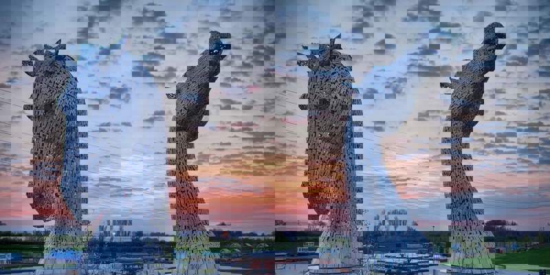 Two massive, sculptural horse heads made of steel rise from the water at sunset in Scotland.