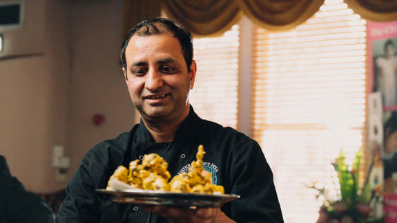  Chef in a black jacket smiling and holding a plate of Indian food.
