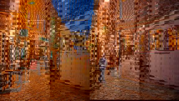 Twinkling fairy lights illuminate Ashton Lane, a charming cobblestone street lined with brick buildings, pubs, and restaurants in Glasgow at dusk.