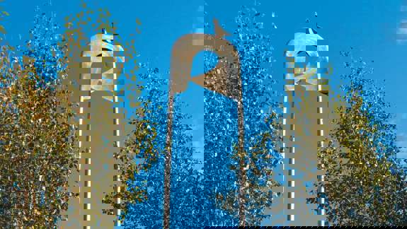 A large, shiny, metallic safety pin sculpture stands tall between two trees against a blue sky.