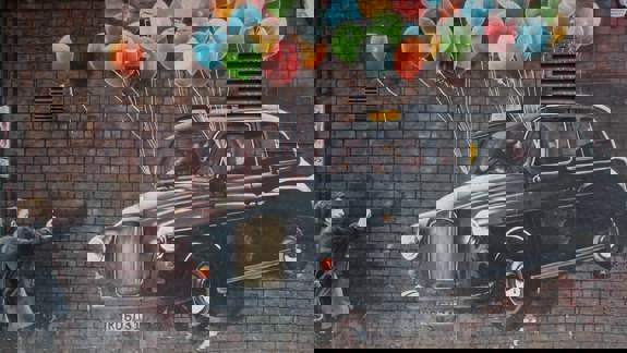 A mural on a brick wall showing a man in a black taxi being lifted into the air by colorful balloons.