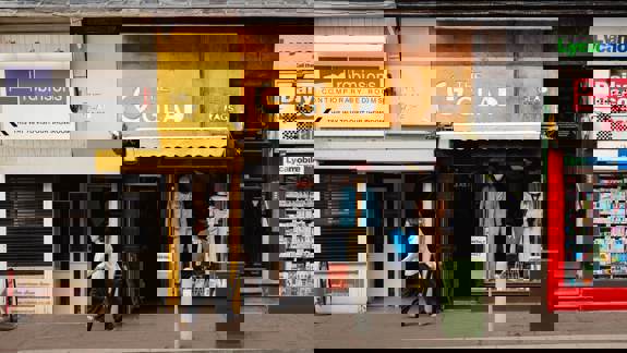 Exterior shot of a city street featuring several shops. The center shop, "The Glad Rags," has a yellow and wood-panelled facade with a window displaying vintage clothing on mannequins. A person is walking past the shop's entrance.