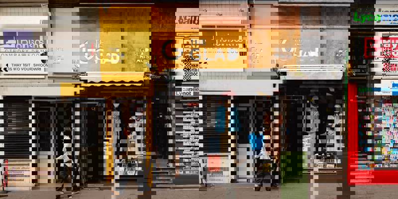 Exterior shot of a city street featuring several shops. The center shop, "The Glad Rags," has a yellow and wood-panelled facade with a window displaying vintage clothing on mannequins. A person is walking past the shop's entrance.