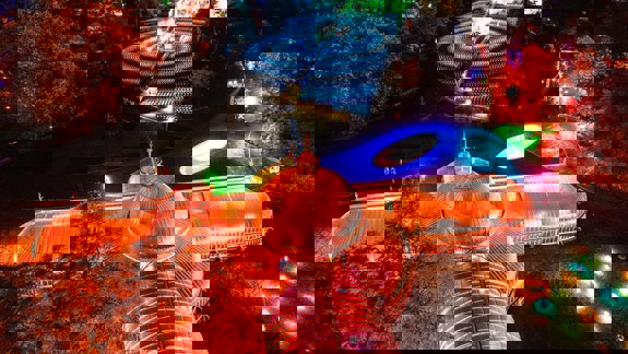 An aerial night view of the illuminated Kibble Palace and surrounding Botanic Gardens, featuring bright orange and pink lights, and a swirling blue light display on the grass.