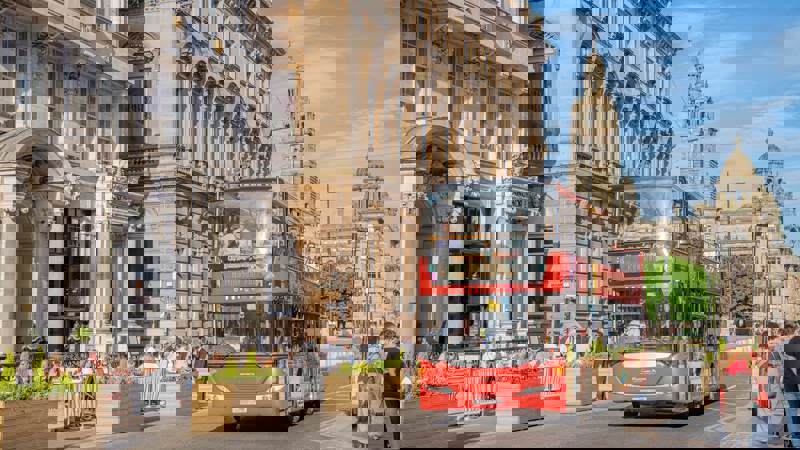 A red open-top tour bus drives past historic buildings in Glasgow on a sunny day, with people dining outdoors and walking nearby.