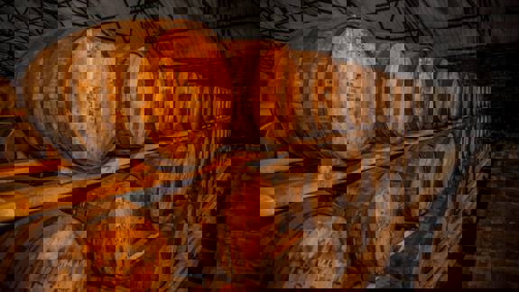 A warehouse with two long rows of wooden barrels filled with whiskey, stacked on shelves. The lighting is dim, highlighting the rich colour of the wood.