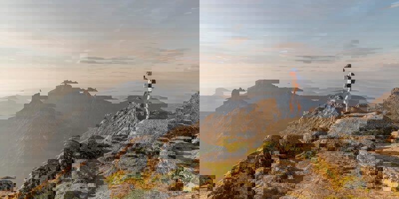 A hiker stands atop the rocky summit of Goat Fell, looking out over the dramatic, jagged peaks and misty glens of the Isle of Arran at sunset.