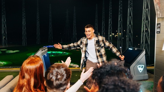 Man holding a golf club celebrating with friends at an indoor driving range at night, with illuminated targets visible in the background.