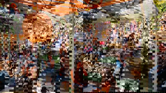 Outdoor seating area with tiered wooden benches under a clear roof, filled with people enjoying drinks