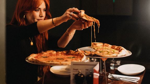 Person pulling a slice of cheesy pizza from a large pizza on a stand, with another pizza and plates on a wooden table in a dimly lit restaurant