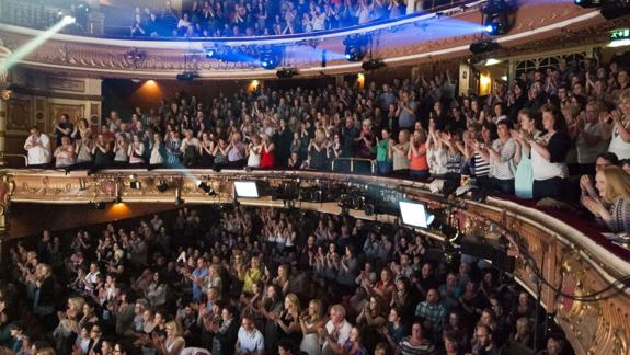 The audience in the grand King's Theatre gives a standing ovation. Ornate balconies, spotlight beams, and a vibrant crowd create an atmosphere of excitement and admiration.