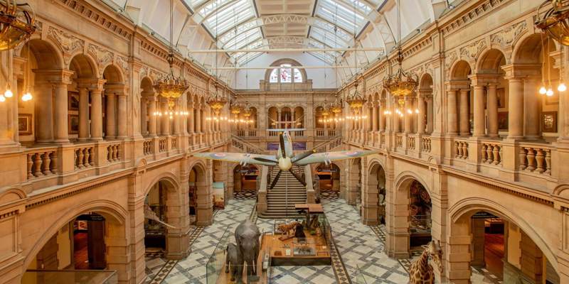 The grand, vaulted interior of a museum with a plane suspended from the ceiling and taxidermy animals, including an elephant and a giraffe, on the floor.