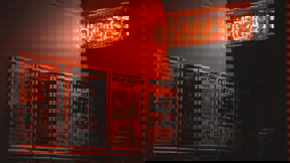  A wall of framed gig posters in a dark space, illuminated by an orange neon light that reads "PIZZAS"