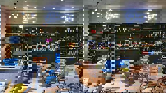 Lounge area at Aparthotel Adagio Glasgow with comfortable seating, shelves stocked with books and snacks, and neon “Library” and “Shop” signs creating a relaxed social atmosphere.