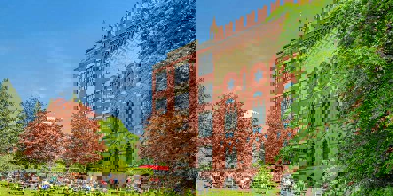 A colourful historic building with ornate windows and people sitting at picnic tables on a grassy lawn.