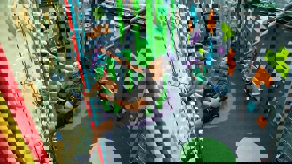 An athletic man is indoor rock climbing with a harness and rope. Another person is climbing on green poles in the background, and another person is on the ground.