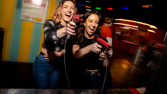 Two people laugh while playing an arcade-style shooting game at Fayre Play in Glasgow, surrounded by colourful lights and carnival-themed décor.