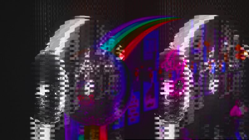 A disco ball hangs in the foreground with a rainbow archway and people in a bar in the background.