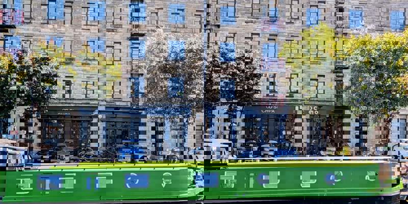 A bright green narrowboat is moored on a canal, with a stone apartment building and the Ocho Spa and café visible on the other side.