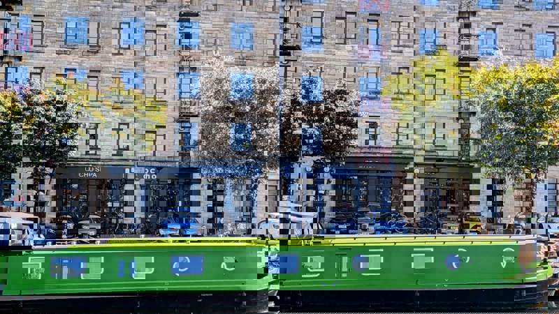 A bright green narrowboat is moored on a canal, with a stone apartment building and the Ocho Spa and café visible on the other side.