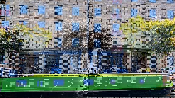 A bright green narrowboat is moored on a canal, with a stone apartment building and the Ocho Spa and café visible on the other side.