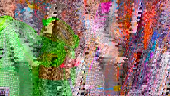 Two individuals wearing protective ponchos are in a room covered in colorful paint splatters, working on their own canvases at a splatter paint studio.