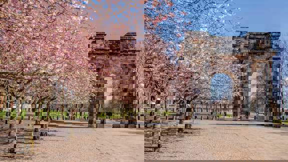 Pink cherry blossoms line a path leading to a large stone arch on a bright spring day.