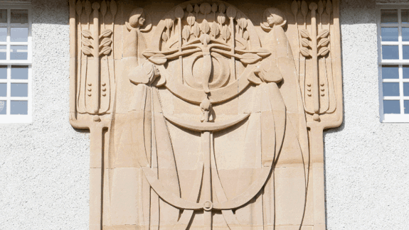 A close-up shot of a stone carving from Charles Rennie Macintosh's House for an Art Lover. Featuring two cloaked figures, with wheat stalks and other decorative elements. It's centered on a light-colored building between two windows.