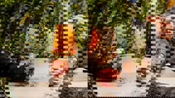 Three hands hold bright orange and minty cocktails on a patio table, surrounded by lush green and yellow foliage.